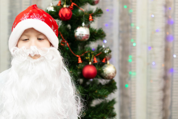 新年前夕，一个7岁的小男孩打扮成留着胡子的圣诞老人，在家里的圣诞树和花环的背景下。有选择性的重点图片下载