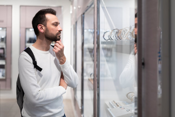 照片中，一个成年英俊的黑发男子身穿白色运动衫，背着双肩包，在购物中心的一家商店里选择了一只时尚的手表图片下载