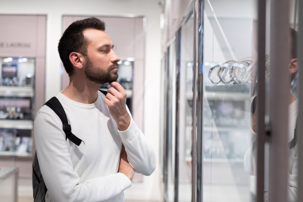 照片中，一个成年英俊的黑发男子身穿白色运动衫，背着双肩包，在购物中心的一家商店里选择了一只时尚的手表图片下载