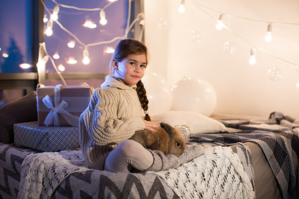 非常漂亮和迷人的黑头发小女孩穿着白色毛衣抱着一只活兔子在房子的内部。新年。圣诞节。野兔。工作室。“r”n图片下载