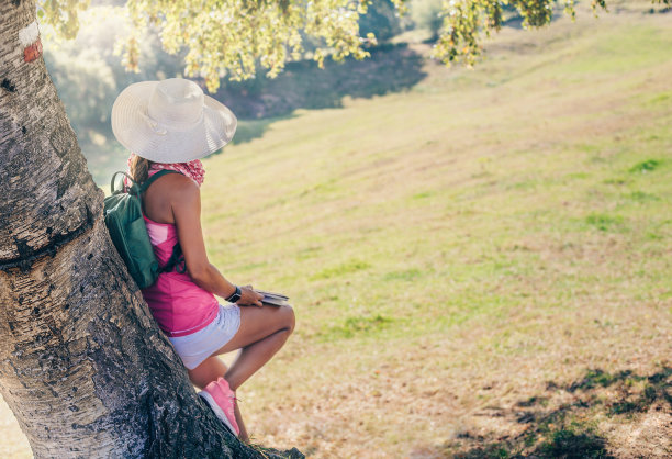 一名戴着白色帽子的女性徒步旅行者倚靠在一棵树上，手里拿着一份议程。她看着被大自然包围的地平线。概念自然、生态和环境。图片下载