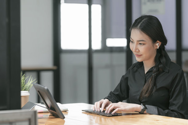 一名妇女坐在桌旁用便携式平板电脑在键盘上打字。图片下载