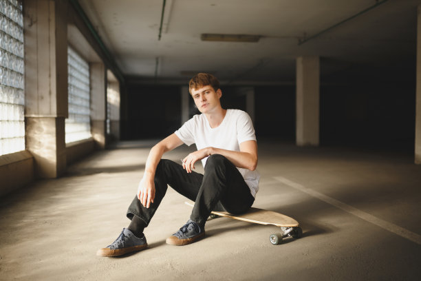 有棕色头发的酷男孩坐在滑板上，幻想地看着镜头吸烟。穿着白色t恤的年轻男子坐在那里，手里拿着香烟，对着镜头摆姿势图片下载