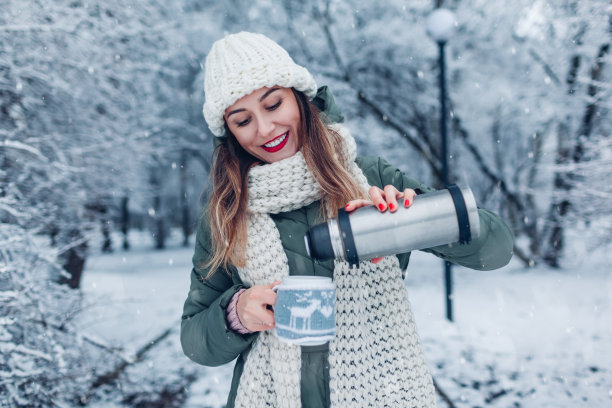 在下雪的冬季公园里，一名女子从保温瓶中倒出热茶。杯与饮料穿着灰色针织圣诞毛衣图片下载
