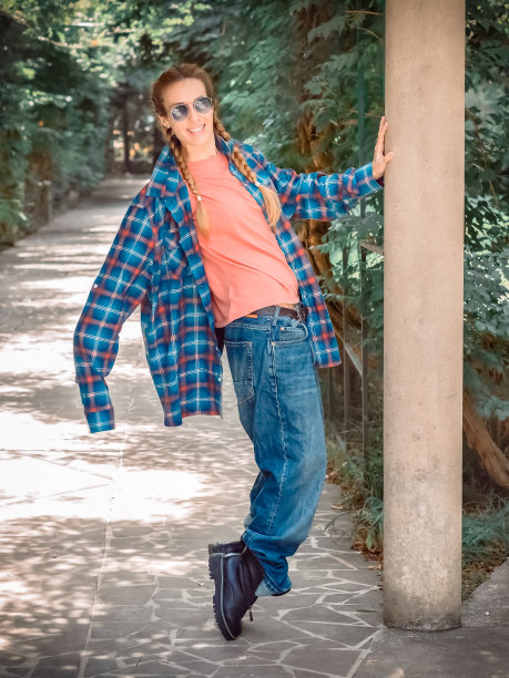 一个女孩穿着长袖子的格子衬衫和巨大的牛仔裤站在绿色小巷的公园里。舞蹈姿势。街舞者图片下载