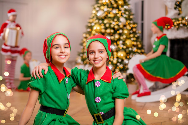 两个欢快快乐的女孩朋友穿着圣诞老人的精灵服装站在圣诞树和其他孩子的背景上图片下载