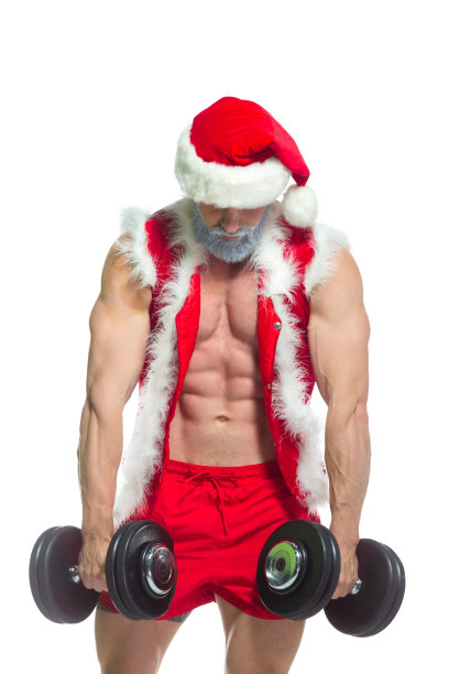 圣诞节。身穿红色短裤、戴着圣诞帽、蓄着灰胡子、肌肉发达的圣诞老人在白色背景的健身房里举着沉重的哑铃图片下载