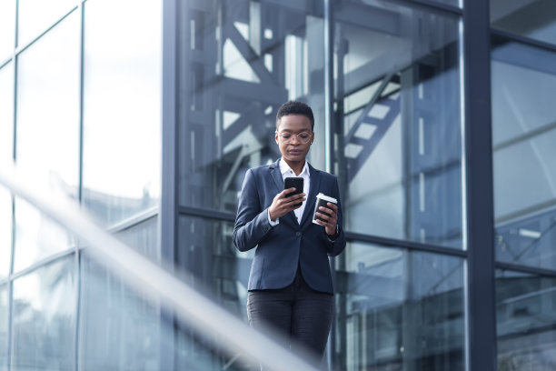 一位自信的非裔美国妇女在商业区走来走去，用着电话。拿着一杯咖啡，穿着正式的衣服。迷人的女商人浏览现代智能手机，同时交流。应用程序图片下载