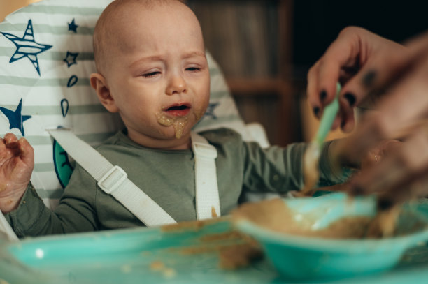 婴儿吃东西时脸上沾满了食物，妈妈用勺子喂他图片下载