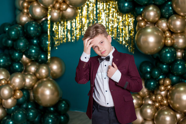 一个小男孩穿着黑色裤子、白色衬衫、领结和紫红色夹克，站在绿色背景上金色和绿色气球组成的拱形中图片下载