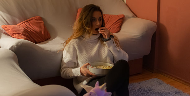 一个漂亮的年轻女人坐在家里的地板和沙发上，在黑暗中看电影，吃爆米花，微笑，享受她的空闲时间和周末活动的高清照片。图片下载