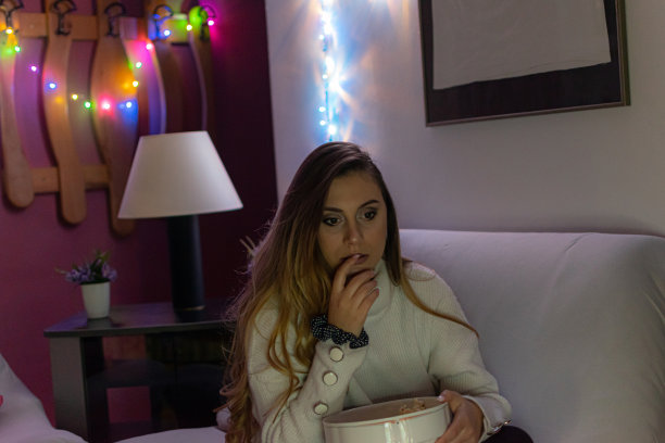 一个漂亮的年轻女人坐在家里的地板和沙发上，在黑暗中看电影，吃爆米花，微笑，享受她的空闲时间和周末活动的高清照片。图片下载