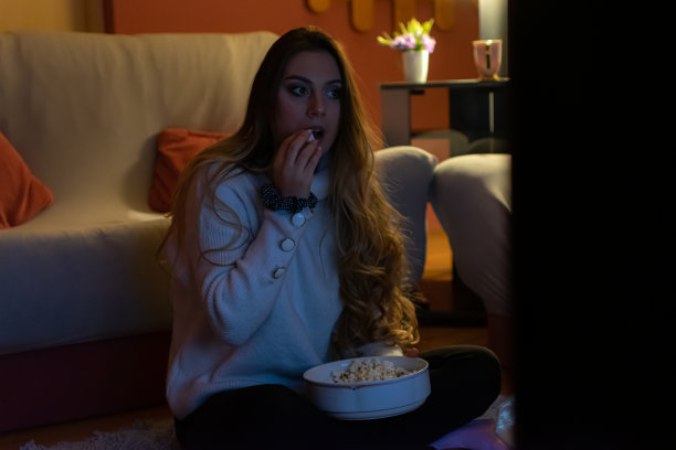 一个漂亮的年轻女人坐在家里的地板和沙发上，在黑暗中看电影，吃爆米花，微笑，享受她的空闲时间和周末活动的高清照片。图片下载