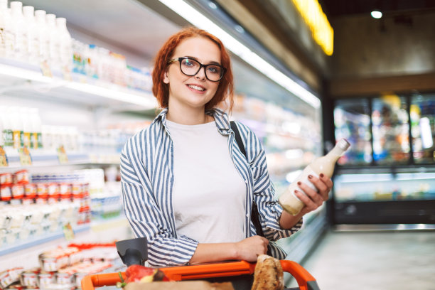 在现代超市里，一个戴着眼镜、穿着条纹衬衫、推着购物车的微笑女孩手里拿着一瓶牛奶，在镜头前快乐地看着图片下载