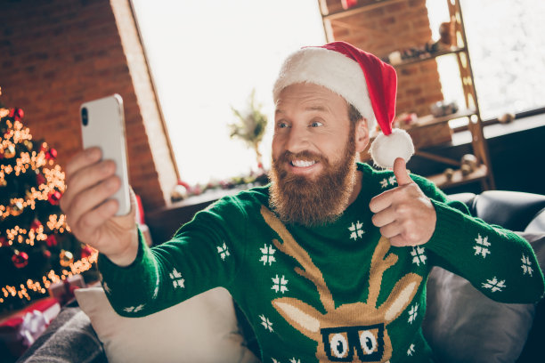 照片不错的家伙拿着手机拍照自拍举起拇指戴圣诞帽套衫装饰家里室内图片下载