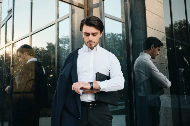 年轻的严肃的长着胡子的深色头发的男人，穿着白色衬衫和经典的夹克，肩上挎着一个袋子，里面装着文件，看着手表，背景是玻璃建筑图片下载