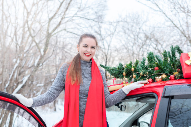在圣诞节和冬季假期期间，美丽的年轻女子站在冬天森林里为圣诞节装饰的红色汽车里图片下载