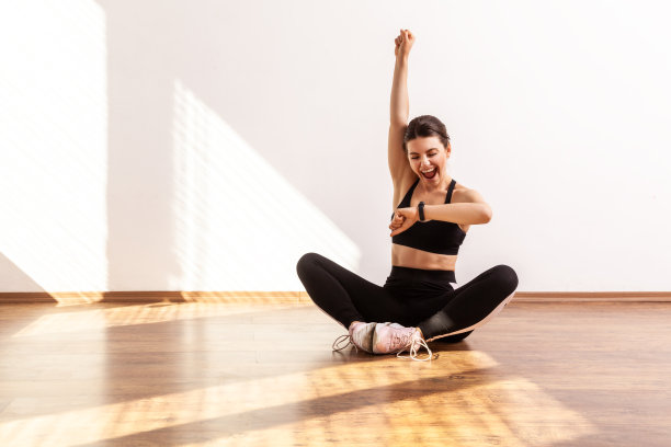 兴奋的运动女孩坐在莲花姿势，看着跟踪器，举起手臂和高兴她的成功图片下载