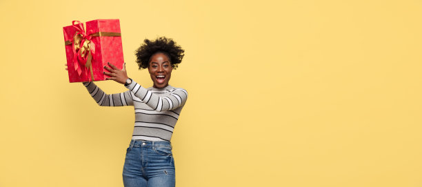 肖像欢快漂亮的非洲女孩在休闲的衣服拿着大礼品盒孤立在黄色的背景。概念的情感，假期图片下载