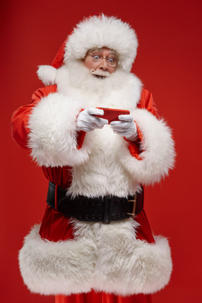 圣诞老人在玩他的红色智能手机，站在红色背景上，他不是在看相机，我们看到他在专业工作室的灯光下图片下载