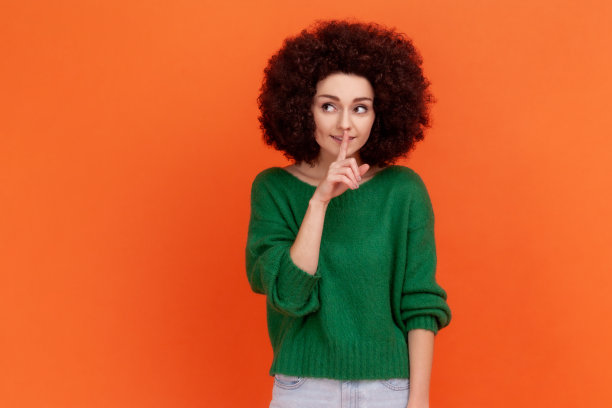 年轻的成年妇女，留着非洲式发型，穿着绿色毛衣，望着别处，手指靠近嘴唇，保守秘密，表达积极的情绪。图片下载
