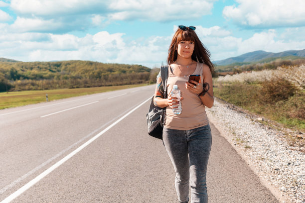 照片上的漂亮年轻女子背着背包，手上有纹身，站在空旷的路上，用着智能手机。副本的空间。本地旅游和在线GPS的概念图片下载