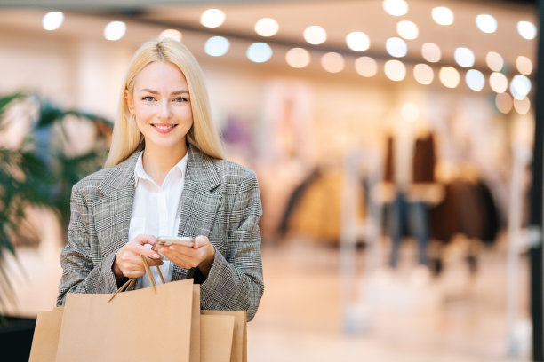 照片的微笑迷人的金发年轻女子在时尚的服装看着相机，拿着手机和购物纸袋与购买在商场。图片下载