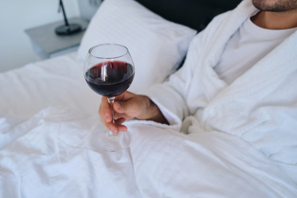 近距离照片年轻迷人的男子在白色浴衣躺在床上与一杯红酒在手现代酒店图片下载