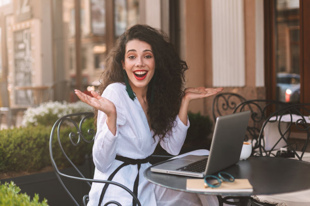 一头黑色卷发，身穿白色服装的微笑女士坐在桌子旁，拿着笔记本电脑，在街上的咖啡馆里愉快地看着相机图片下载