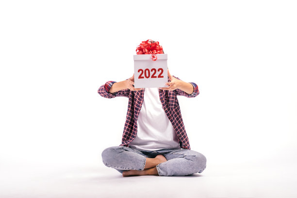 白色背景的年轻女子用写着“2022”的白色礼品盒遮住了自己的脸。这个女孩穿着牛仔裤、衬衫和白色t恤。2022年新年的概念。图片下载