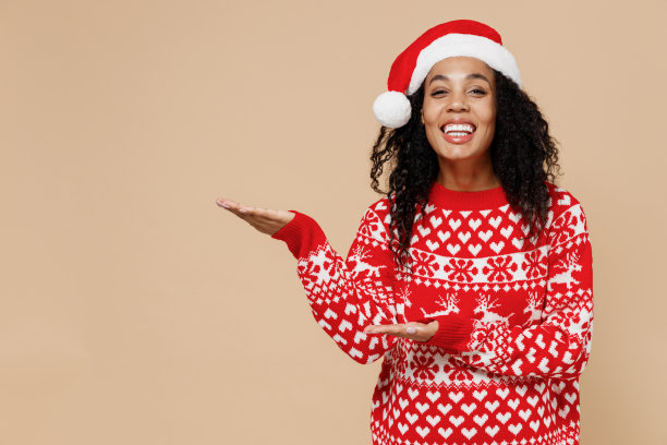 微笑的年轻的美国圣诞老人妇女在圣诞帽毛衣点在空的手掌与复制空间的地方模拟孤立在柔和的米色背景工作室肖像。新年快乐圣诞假期概念。图片下载