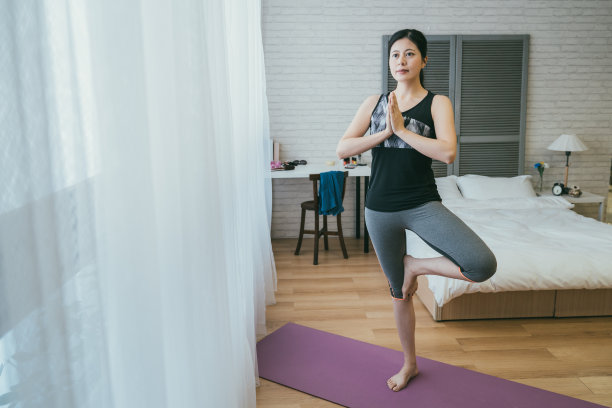 在明亮的家庭卧室室内晨练期间，完整长度的亚洲运动女人是把手掌放在一起，在瑜伽垫上做树的姿势。图片下载