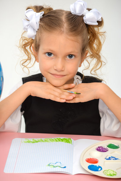 快乐的小女孩坐在桌旁，拿着铅笔和课本。快乐的孩子小学生在桌旁做作业。美丽的微笑少女图片下载