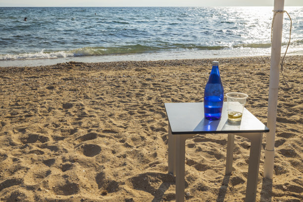 看到一瓶水站在一个太阳伞下的桌子上的海滩靠近水。希腊。图片下载