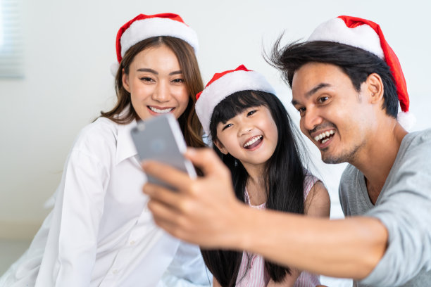 亚洲父母与小女儿视频电话在线与家人。可爱的爸爸、妈妈和小女孩坐在卧室的床上，周末在家里用手机和亲戚聊天。图片下载