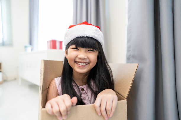 亚洲小女孩戴圣诞帽，坐在礼品盒里。可爱的小孩子兴奋而快乐地庆祝节日圣诞感恩节派对，在包裹里玩耍，看着相机。素材图片