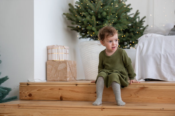 小女孩穿着温暖的绿色连衣裙坐在圣诞树旁的木台阶上，阁楼里有花环图片下载