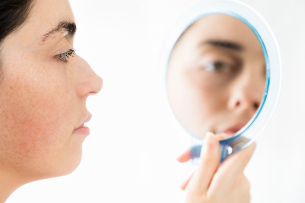 鼻子整形手术。一个年轻的白人妇女的近距离脸看着她的座头鲸鼻子在镜子里，孤立在一个白色的背景。鼻整形术的概念图片下载