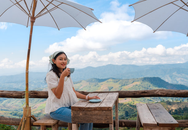 在泰国清莱的旅游景点土井昌，一名亚洲女子放松地坐着，拿着咖啡杯在露台上的木桌上，带着白色的伞，俯瞰着山景和多云的蓝天图片下载