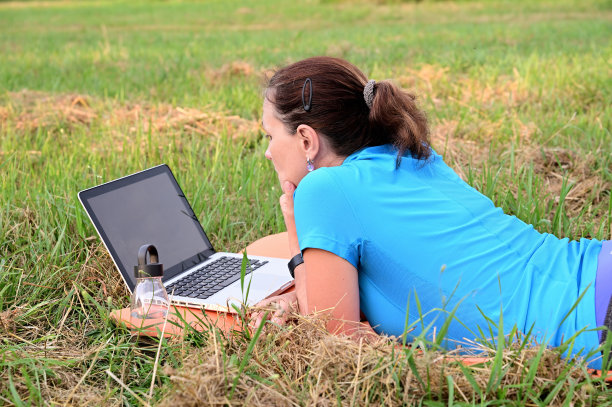 一名女子从笔记本电脑屏幕上阅读新闻。她趴在公园的运动垫子上图片下载