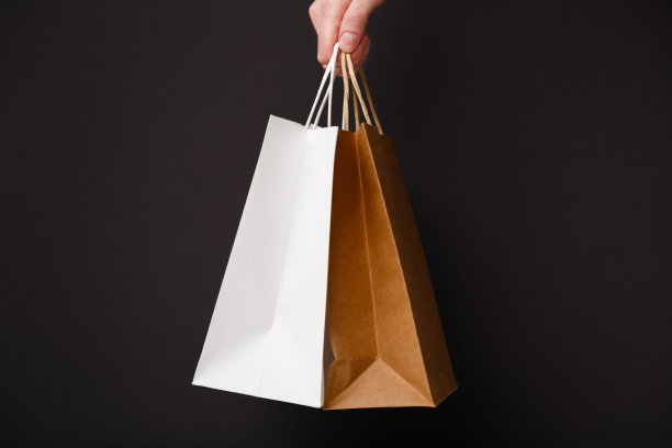 女性手拿两个购物袋孤立在黑色背景上。手里拿着白色和棕色的工艺空白纸袋。黑色星期五大甩卖、打折、购物、生态理念图片下载