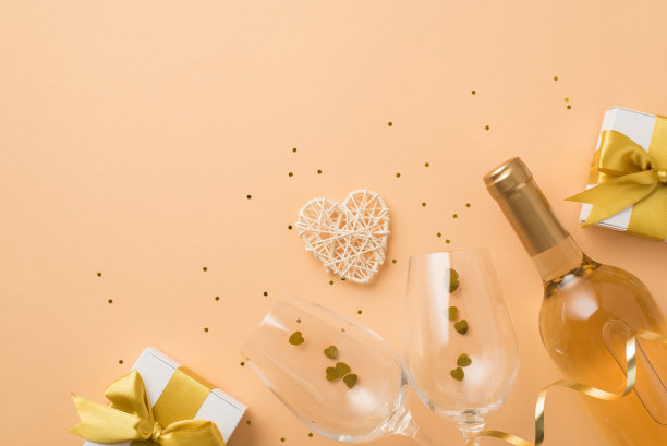 顶视图照片的情人节装饰白色礼品盒与金色蝴蝶结葡萄酒酒瓶藤心葡萄酒杯与心形的纸屑和亮片在孤立的柔和的米色背景与copyspace图片下载