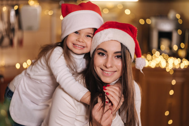 圣诞节期间，可爱的小女儿和妈妈在厨房里的照片。戴圣诞帽的女孩。装饰适合树在桌子上。女孩做姜饼图片下载