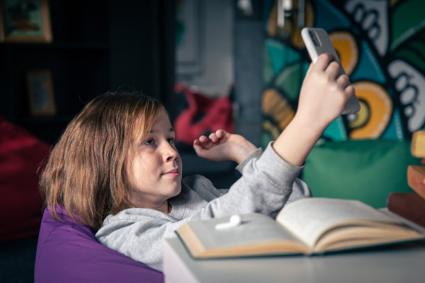 一个十几岁的女孩用智能手机自拍，而不是看书。图片下载