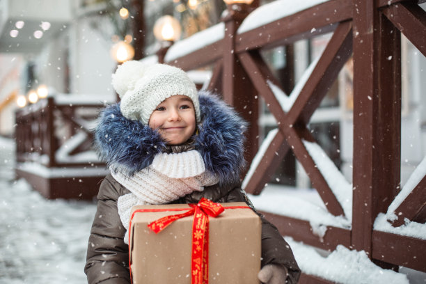 这张照片拍摄的是一个快乐的女孩，她拿着圣诞礼盒站在冬天下雪的城市街道上，节日市场上到处都是装饰品和彩灯。保暖衣物、针织帽、围巾及毛皮。新的一年图片下载