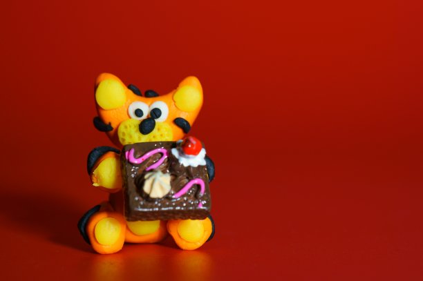 一只橡皮泥做的玩具老虎，红色的背景上，手里拿着一个蛋糕。图片下载