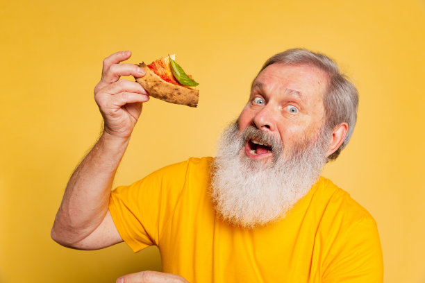 情绪激动的灰色胡子男人在黄色的工作室背景下品尝奶酪和番茄披萨。图片下载