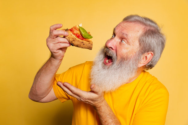 激动的灰胡子男人吃着美味的披萨，奶酪和西红柿被隔离在黄色的画室背景上。图片下载