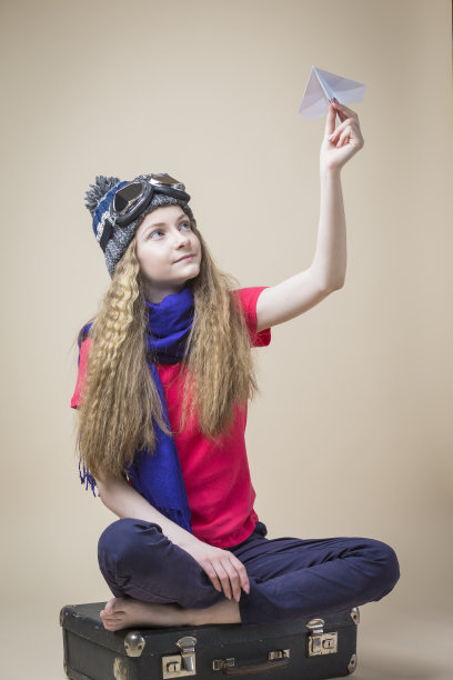 十几岁的女孩在飞行的眼镜和针织温暖的帽子与折纸飞机摆姿势，而坐在旧手提箱米色的背景。图片下载