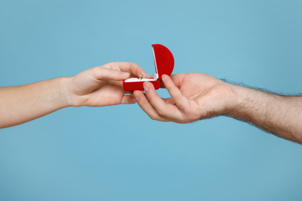 近距离裁剪照片的女性男性手握红色的盒子与婚戒求婚孤立在柔和的蓝色背景在工作室。情人节假期的概念。图片下载